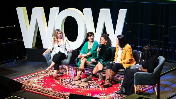 Five women sit on stage in conversation, framed by large "WOW" letters. They appear engaged and cheerful, seated on chairs over a patterned rug.
