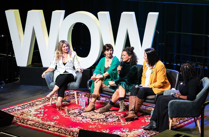 Five women sit on stage in conversation, framed by large "WOW" letters. They appear engaged and cheerful, seated on chairs over a patterned rug.
