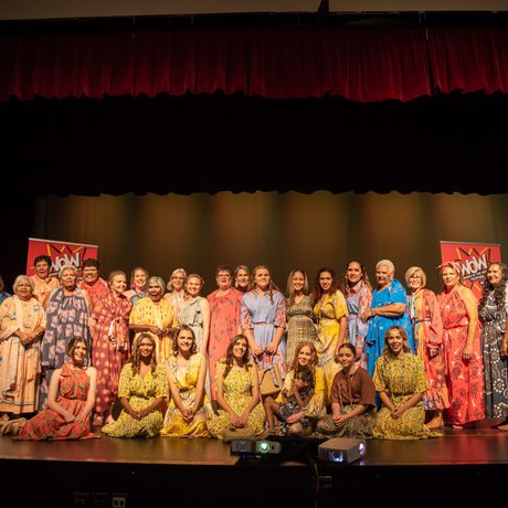 A group of performers in colourful costumes pose on stage with a backdrop under red curtains