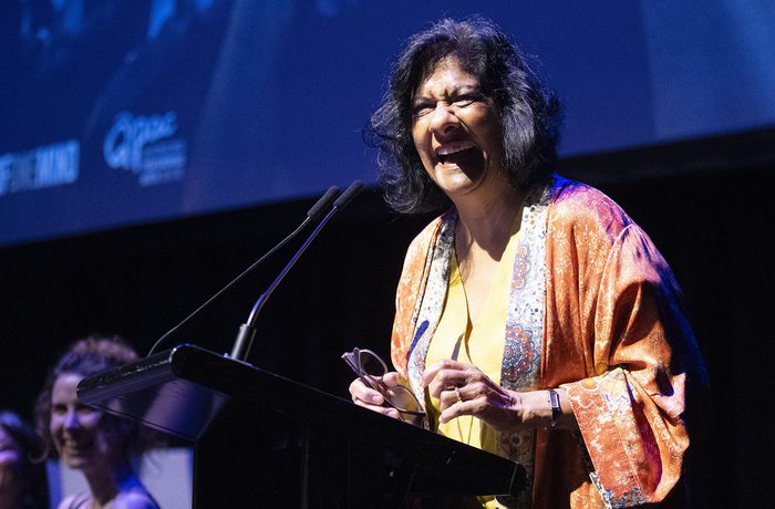 A woman in colourful attire smiles while speaking at a podium on a dimly lit stage, with two people visible in the background, conveying a joyful atmosphere.