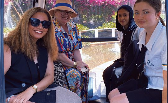 Four people sit inside a cable car, with bright pink flowers visible through the windows. The mood is relaxed and cheerful