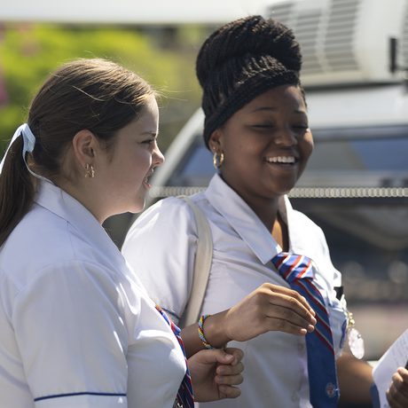 Two women in white uniforms stand outside, smiling and talking on a bright, sunny day