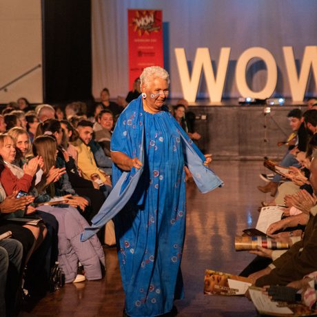 Elderly woman in a vibrant blue sari walks joyfully on a runway, surrounded by applauding audience. "WOW" sign in the background.