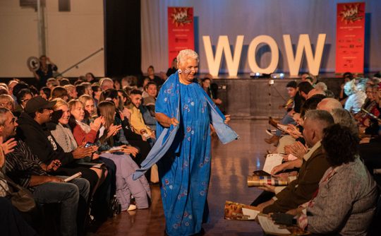 Elderly woman in a vibrant blue sari walks joyfully on a runway, surrounded by applauding audience. "WOW" sign in the background.