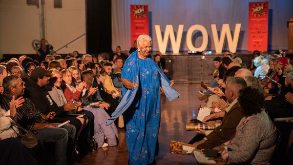 Elderly woman in a vibrant blue sari walks joyfully on a runway, surrounded by applauding audience. "WOW" sign in the background.
