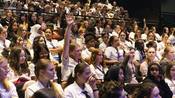 A group of students in a lecture hall, with one raising her hand. The atmosphere is attentive and engaged
