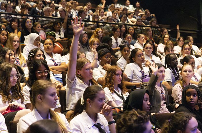 A group of students in a lecture hall, with one raising her hand. The atmosphere is attentive and engaged