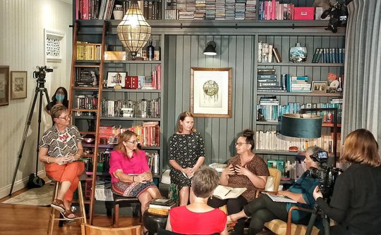 Cozy library room with bookshelves filled with books and decor. Seven people sit in discussion, creating an engaged, warm, and collaborative atmosphere.