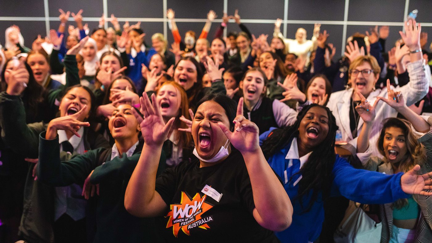 A large, joyful group of diverse students cheer enthusiastically in a school setting. Many wear uniforms, conveying a lively atmosphere.