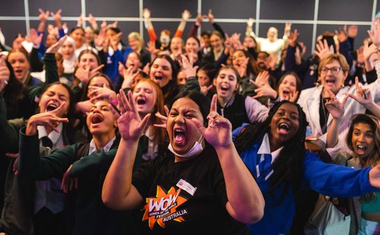 A large, joyful group of diverse students cheer enthusiastically in a school setting. Many wear uniforms, conveying a lively atmosphere.