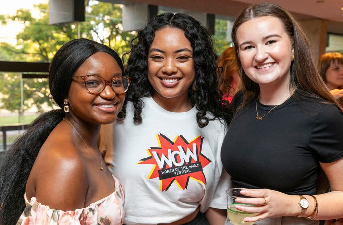 Three women are smiling and standing close together at an indoor gathering. One wears a "WOW" shirt, another holds a drink. The mood is cheerful and friendly.