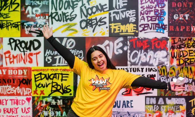 A woman in a bright yellow shirt joyfully poses with arms outstretched in front of a vibrant graffiti wall featuring feminist slogans and colourful art.