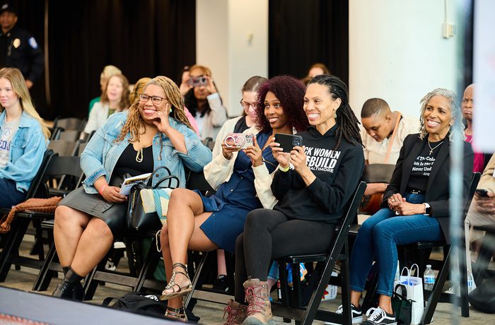 A group of six women sitting in chairs, smiling and engaging at a public event. They hold phones, suggesting they are recording or taking photos.