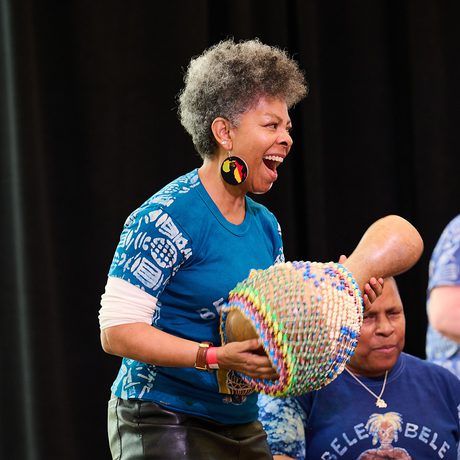 A woman passionately shakes a beaded instrument on stage, wearing a blue patterned shirt.