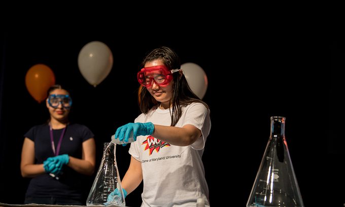 Two people in safety goggles and gloves perform a science experiment on stage, with balloons in the background.