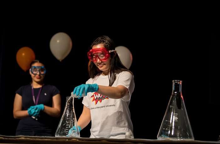 Two people in safety goggles and gloves perform a science experiment on stage, with balloons in the background.