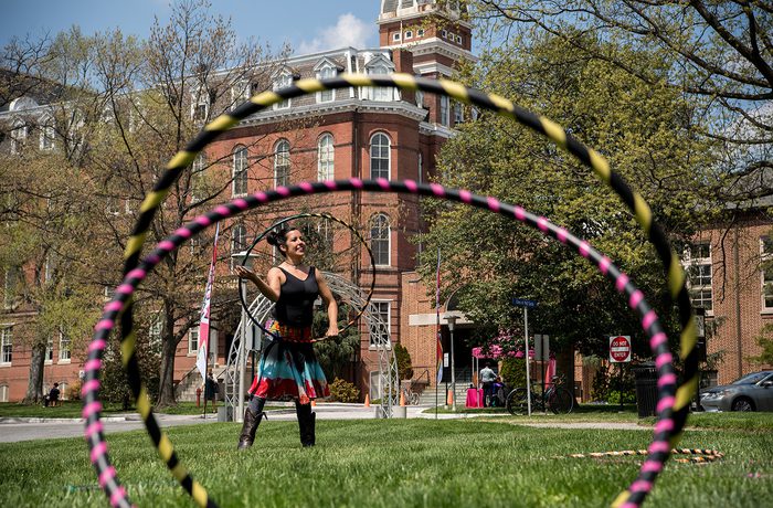 A person hula-hoops on a grassy lawn in front of a historic brick building. The sky is clear and trees frame the scene.