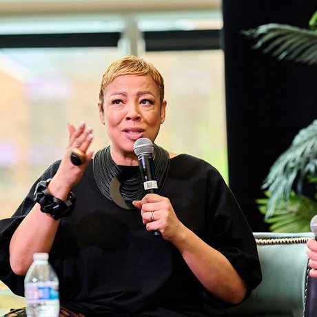 A person in a black outfit, holding a microphone, speaks passionately during an indoor panel event.