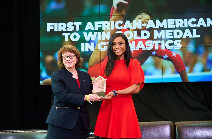 Two women stand in front of a large screen displaying "First African American to win gold medal in Gymnastics." One is presenting an award to the other.