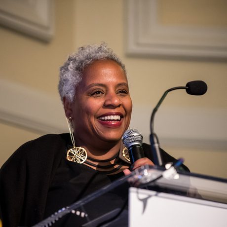 An individual with short grey hair smiles warmly while speaking into a microphone at a podium, conveying a confident and uplifting tone.