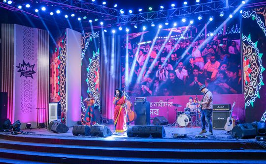 A vibrant stage is lit with blue and pink lights, showcasing a band performing before a lively crowd. A large screen displays audience reactions.