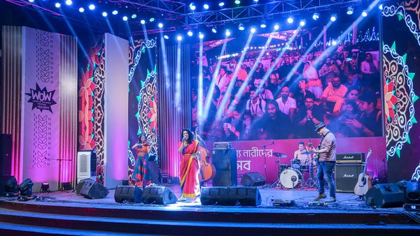 A vibrant stage is lit with blue and pink lights, showcasing a band performing before a lively crowd. A large screen displays audience reactions.