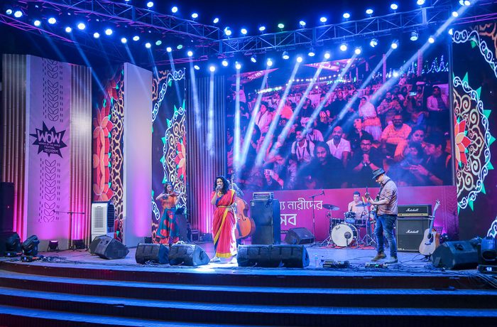 A vibrant stage is lit with blue and pink lights, showcasing a band performing before a lively crowd. A large screen displays audience reactions.