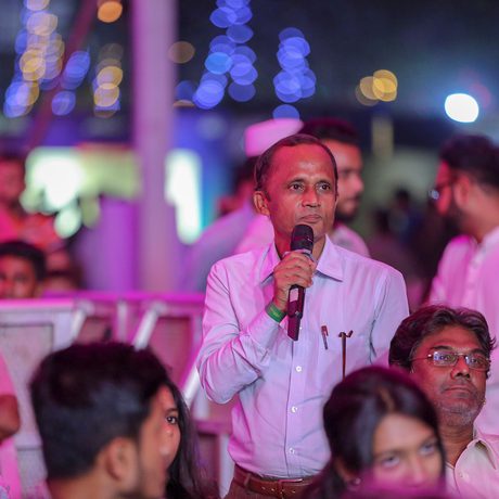 A man in a white shirt speaks into a microphone in a vibrant, well-lit event. People sit around him, engaging attentively.
