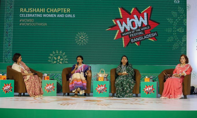 Four women sit in armchairs on a stage, engaging in a panel discussion. The backdrop displays "WOW" and "Celebrating Women and Girls," creating an empowering tone.
