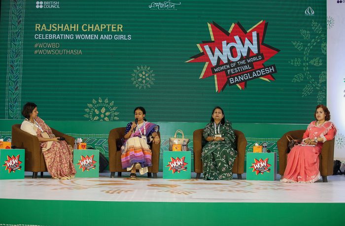 Four women sit in armchairs on a stage, engaging in a panel discussion. The backdrop displays "WOW" and "Celebrating Women and Girls," creating an empowering tone.