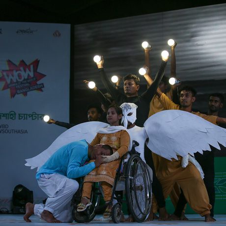 Performers on stage; one in a wheelchair with wings and mask, others surrounding with raised arms holding glowing orbs.