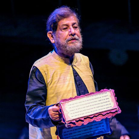 A man with a beard holds a framed certificate on stage. He wears a yellow and black outfit, and the scene is lit with a blue spotlight.