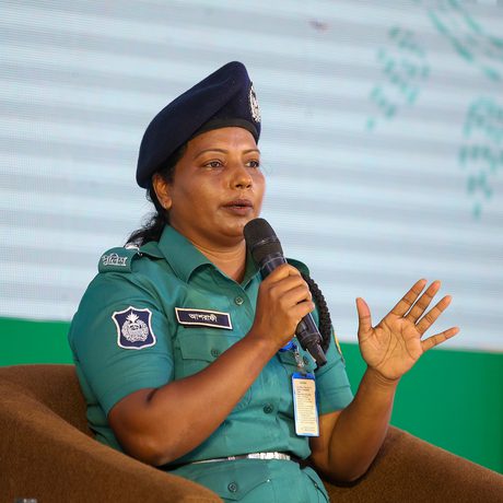 A woman in a teal police uniform and beret speaks into a microphone while seated.