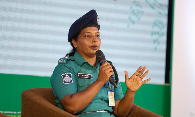 A woman in a teal police uniform and beret speaks into a microphone while seated.