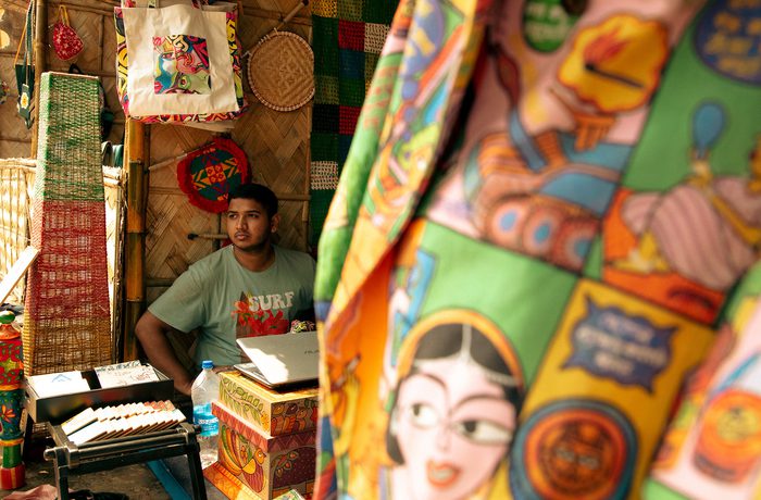 A vendor sits in a colourful market stall surrounded by vibrant textiles showcasing bold, graphic designs.