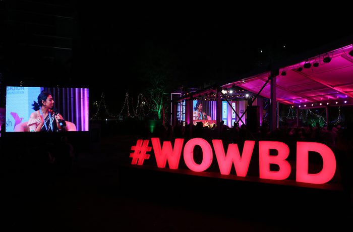 Large illuminated #WOWBD sign glows in pink under a night sky at an outdoor event. A nearby screen shows a person speaking.
