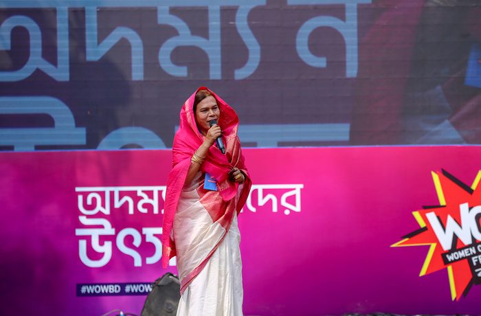 A woman in a white and pink sari speaks onstage, holding a microphone. Behind her is a colourful "WOW" event banner with vibrant pink and blue colours.