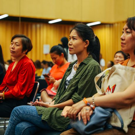A diverse group of focused women sit in a conference room, listening attentively. They wear casual attire, with yellow walls and soft lighting in the background.