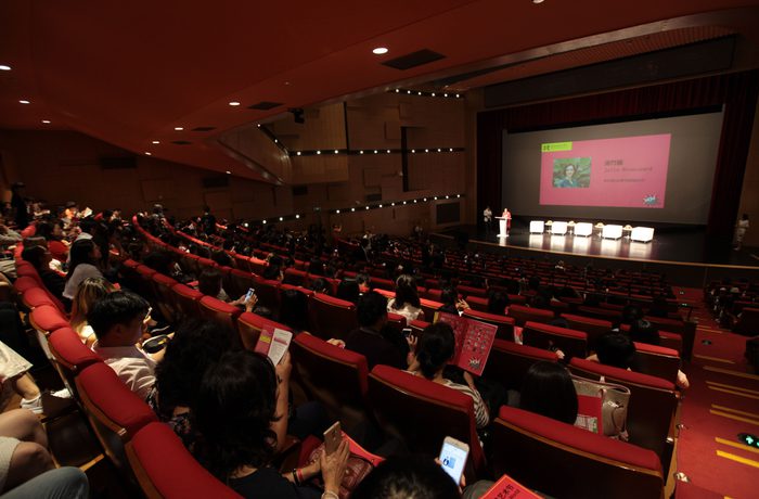 A spacious auditorium with red seats and a crowd facing a stage. A large screen displays a presentation, creating a focused and engaged atmosphere.