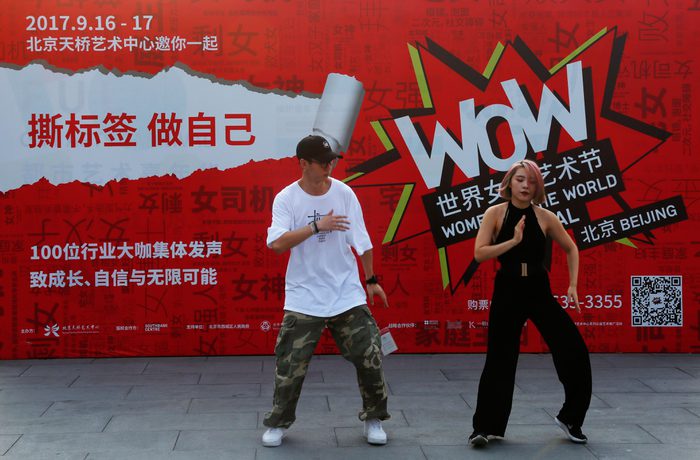 Two people are dancing energetically in front of a vibrant red banner for the "Women of the World" festival in Beijing.