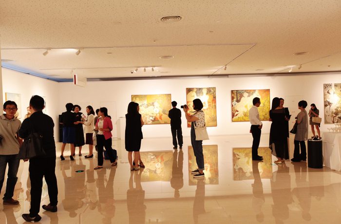 People in a brightly lit art gallery observe paintings on white walls. The polished floor reflects artwork and visitors.