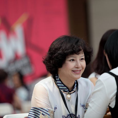 A woman with curly hair smiles warmly while talking to another person. They're sitting indoors with a blurred red "WOW" background