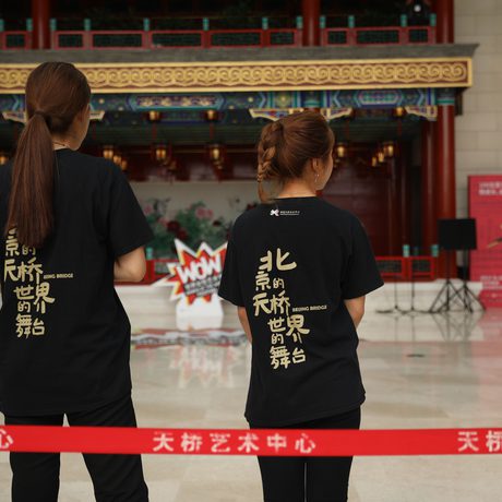 Two people in black shirts with Chinese text on the back stand facing a stage with a "WOW" poster. A red barrier rope is in the foreground.
