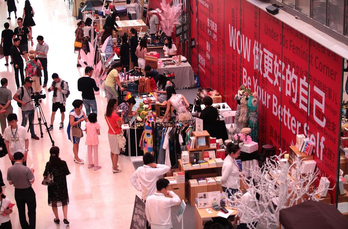 A bustling indoor market features various stalls with colourful clothes and items. Shoppers engage with vendors. A large red banner reads, "Be Your Better Self."