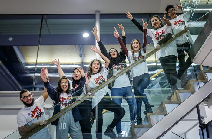 A group of eight people wearing matching white t-shirts printed with the WOW logo eagerly pose on a staircase, waving and smiling