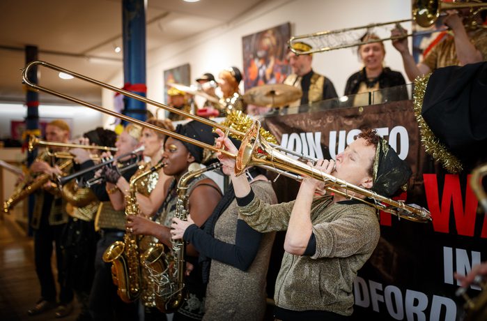 A lively brass band performs indoors, members playing trombones and saxophones.