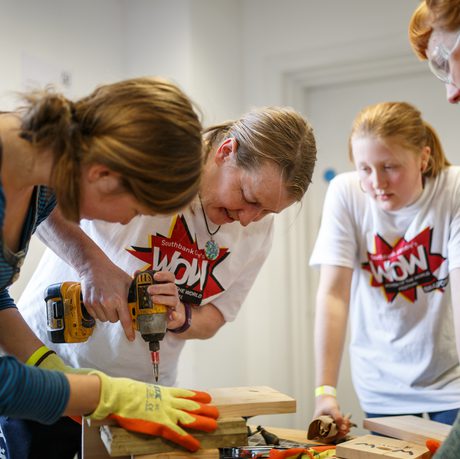Three women in casual clothing focus on a woodworking project. One woman uses a drill, while others watch attentively.