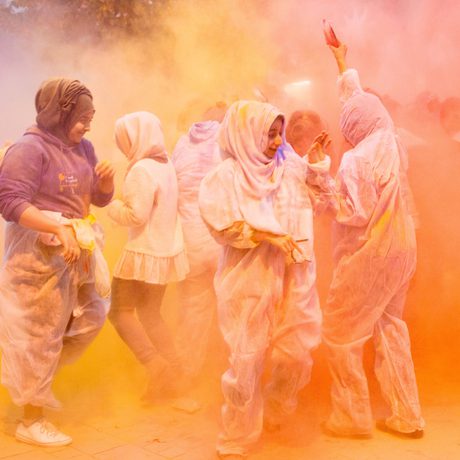 People in white outfits and headscarves are joyfully covered in vibrant orange and yellow powder during a colourful festival.