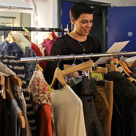 A person smiling while reading a sheet of paper stands beside a clothing rack filled with various outfits in a brightly lit room.