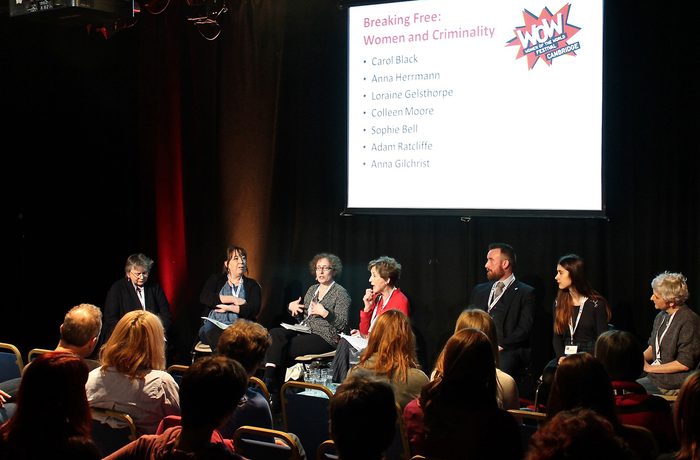 A panel of seven people are seated on stage discussing branding in front of an audience. A screen displays speaker names.